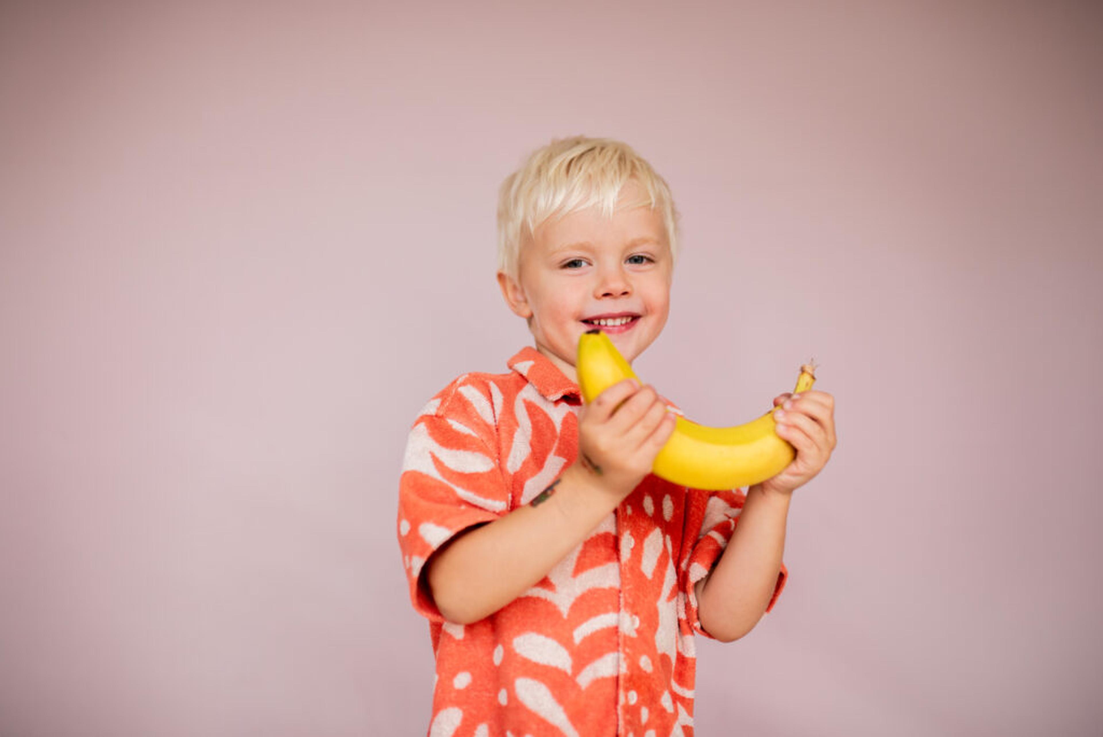 Leende blond pojke i orange mönstrad skjorta som håller i en gul banan
