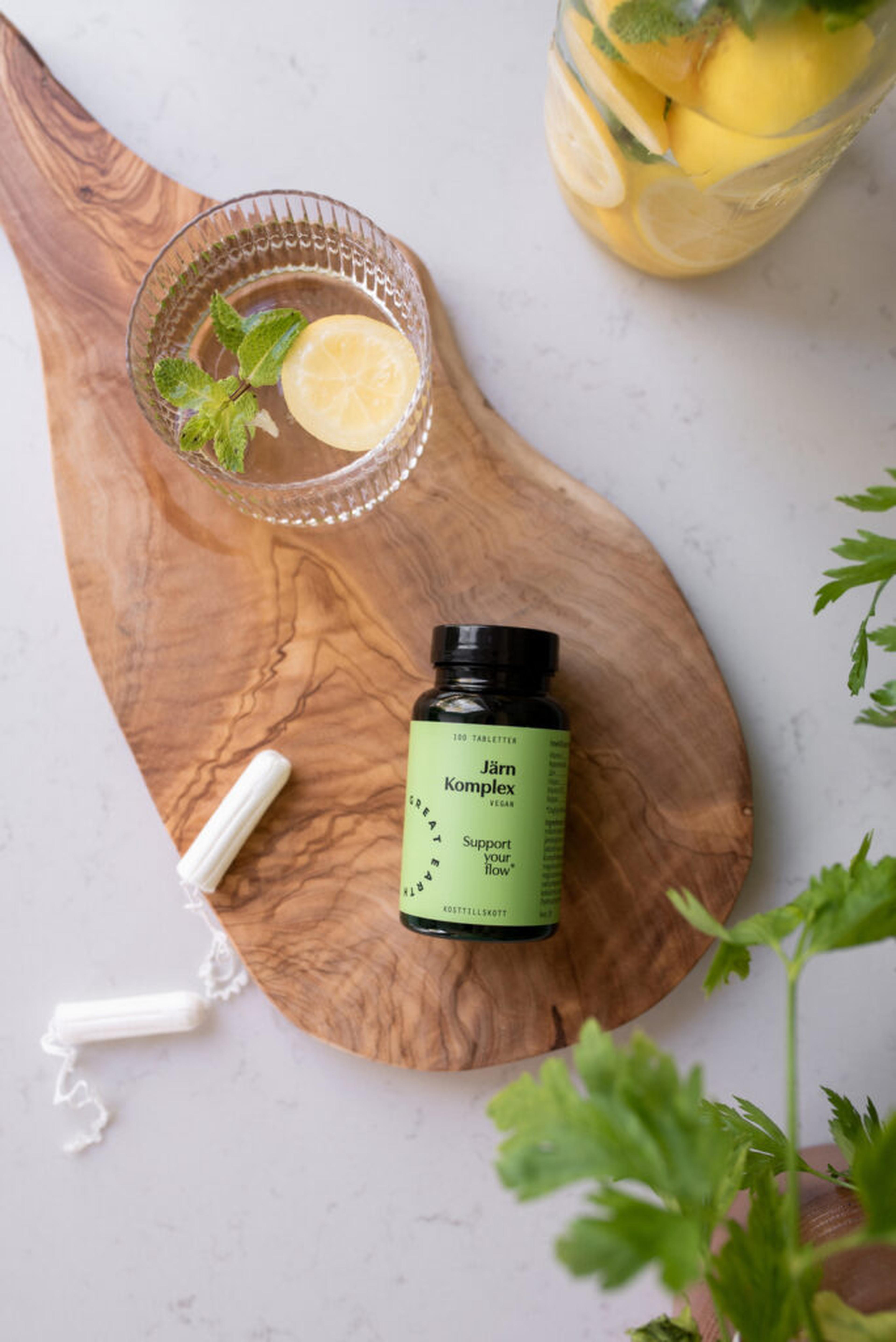 Great Earth Iron Complex bottle beside tampons and a glass of lemon water on a wooden board