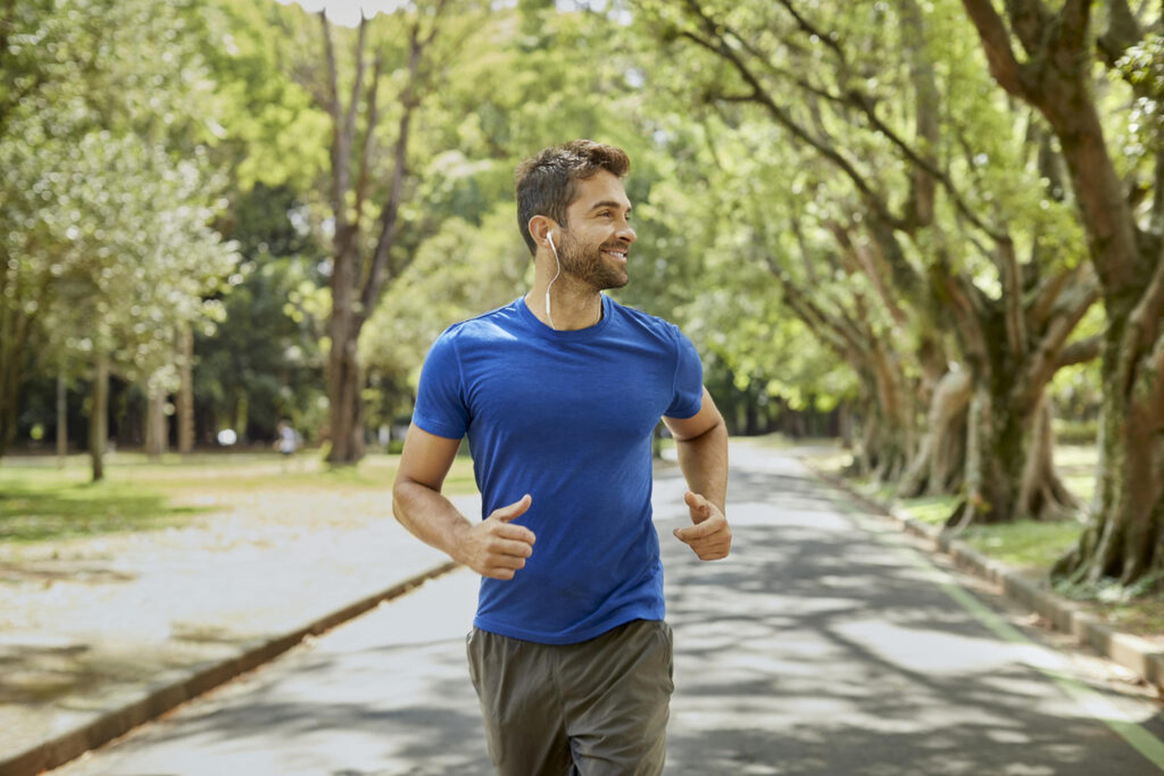 Leende man i blå t-shirt som joggar med hörlurar längs en trädallé i en park