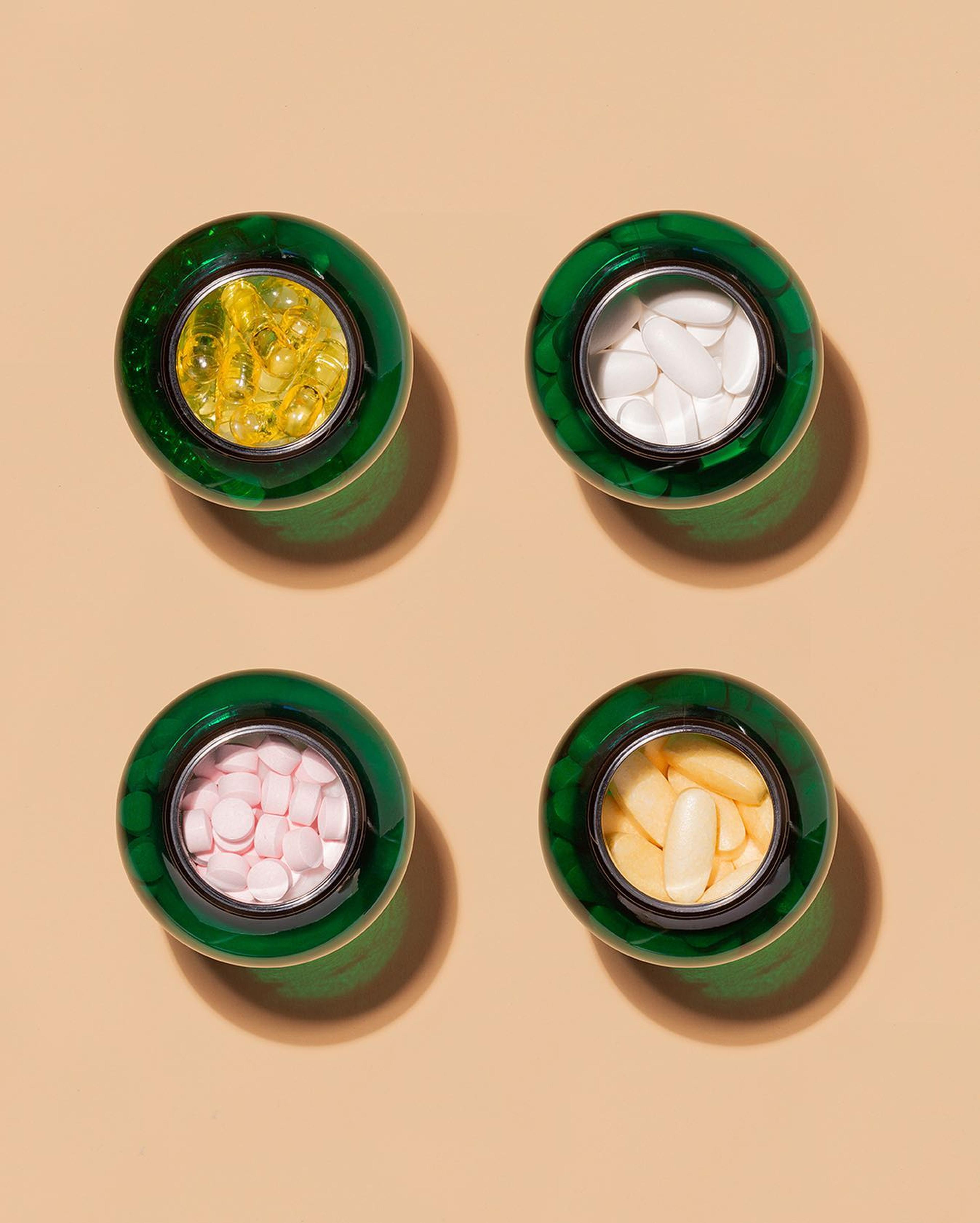 Top view of four open green supplement jars filled with different capsules and tablets on a beige background