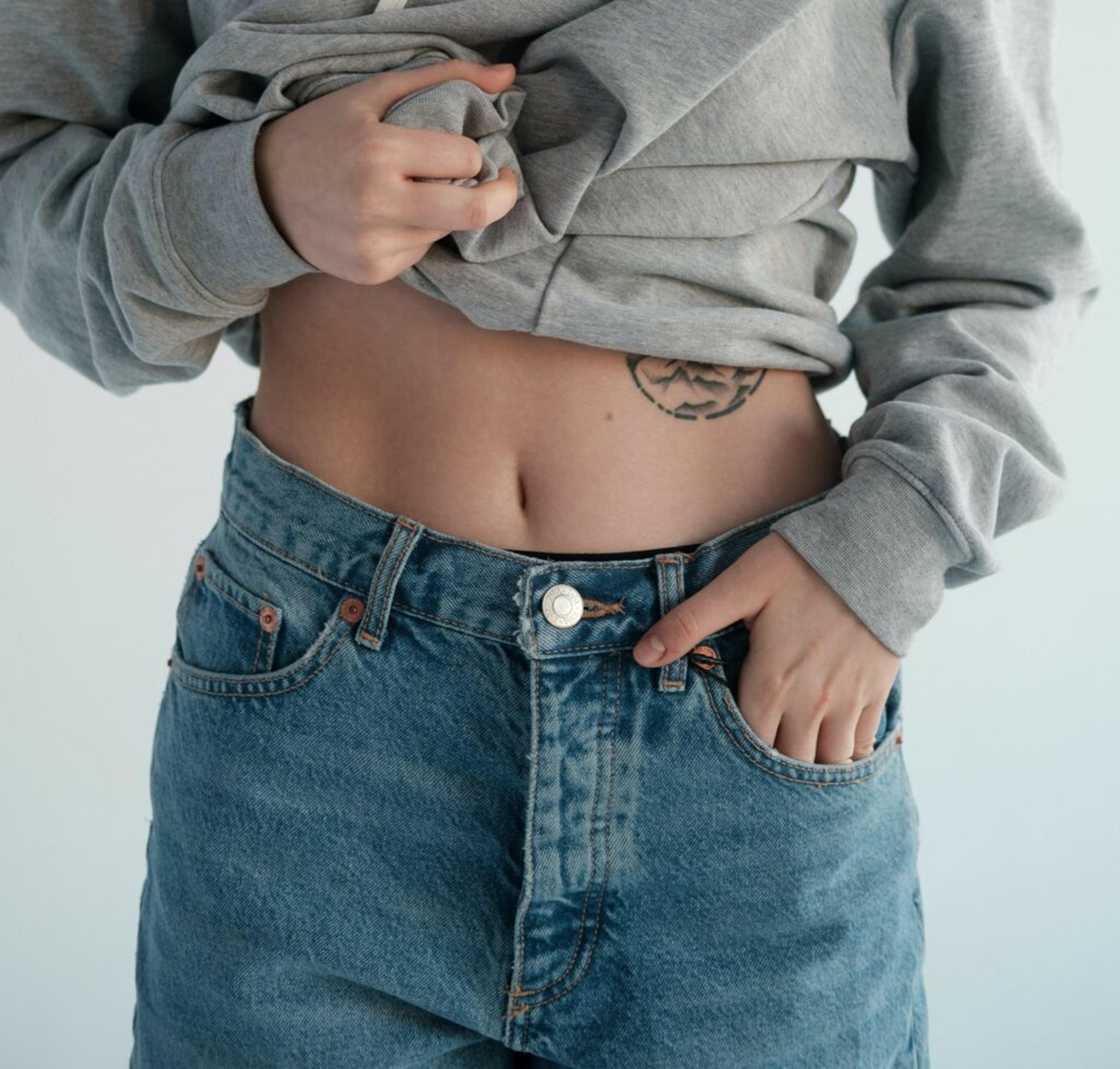 Person som lyfter en grå tröja och visar sin bara mage, klädd i blå jeans