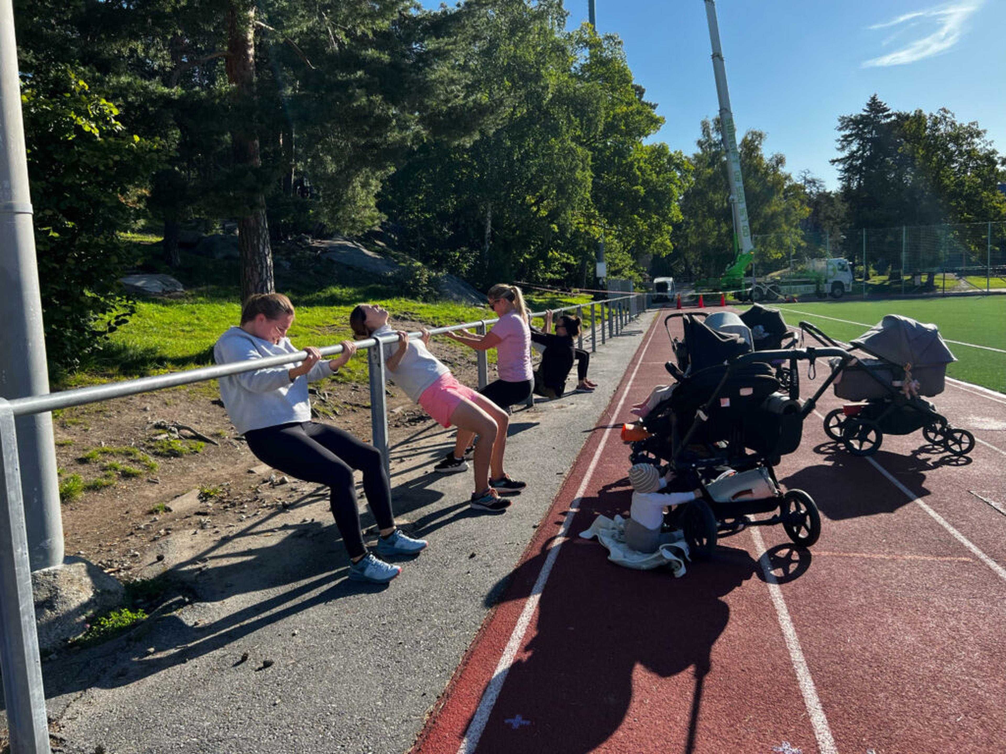 Women doing strength exercises against a railing beside baby strollers on an outdoor running track