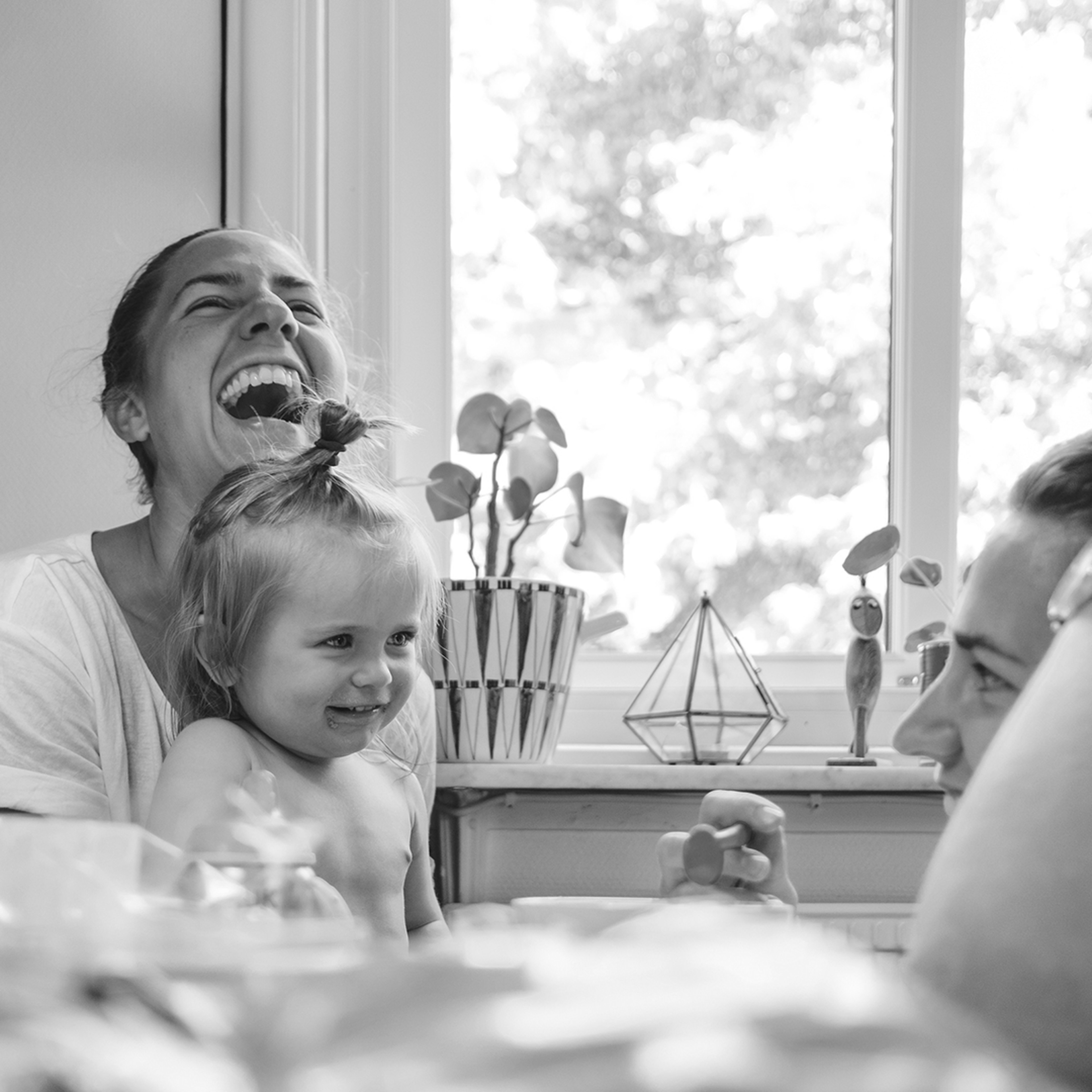 Smiling toddler and laughing woman sitting together at a table by a window