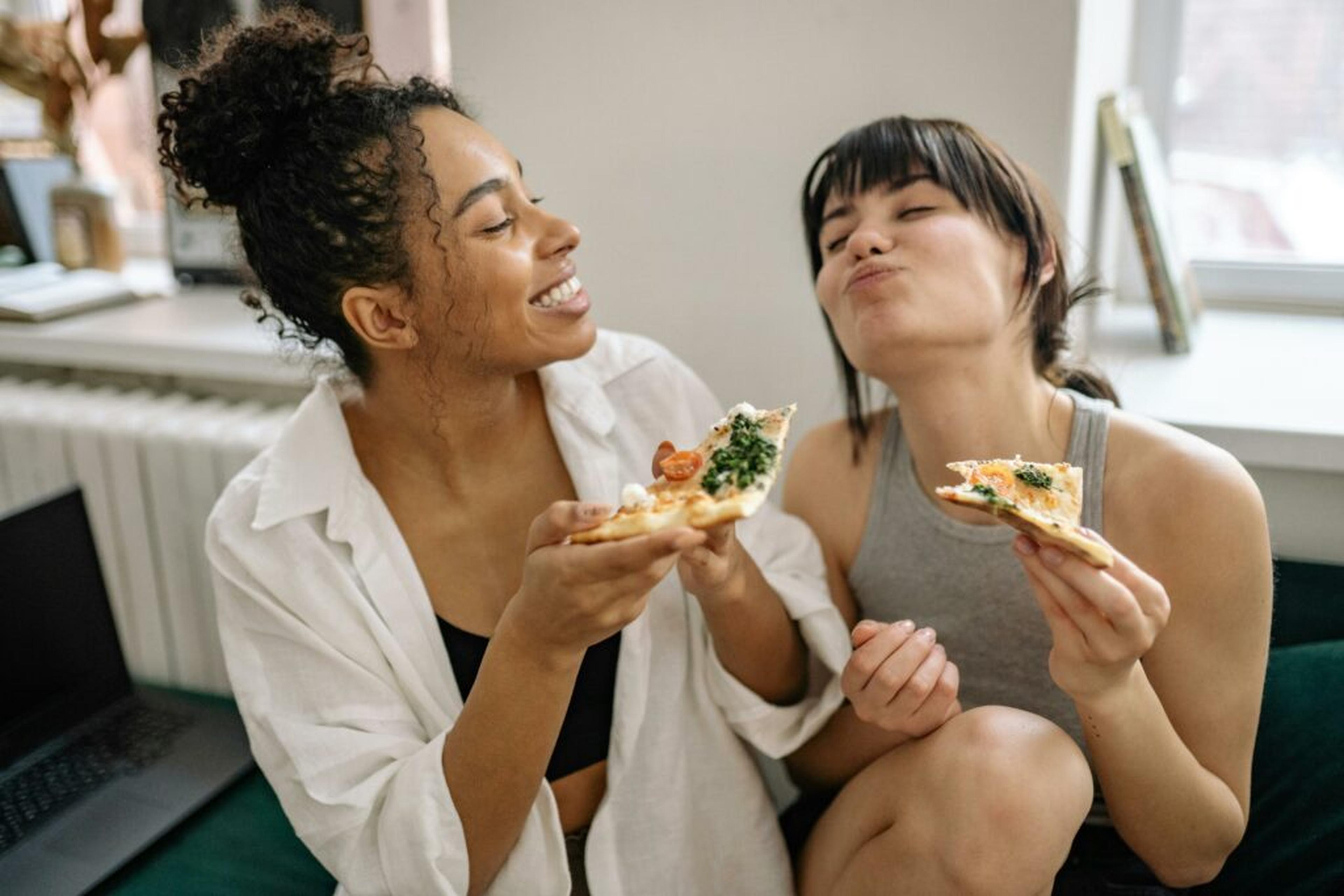 Två leende unga kvinnor som sitter tillsammans och äter pizza