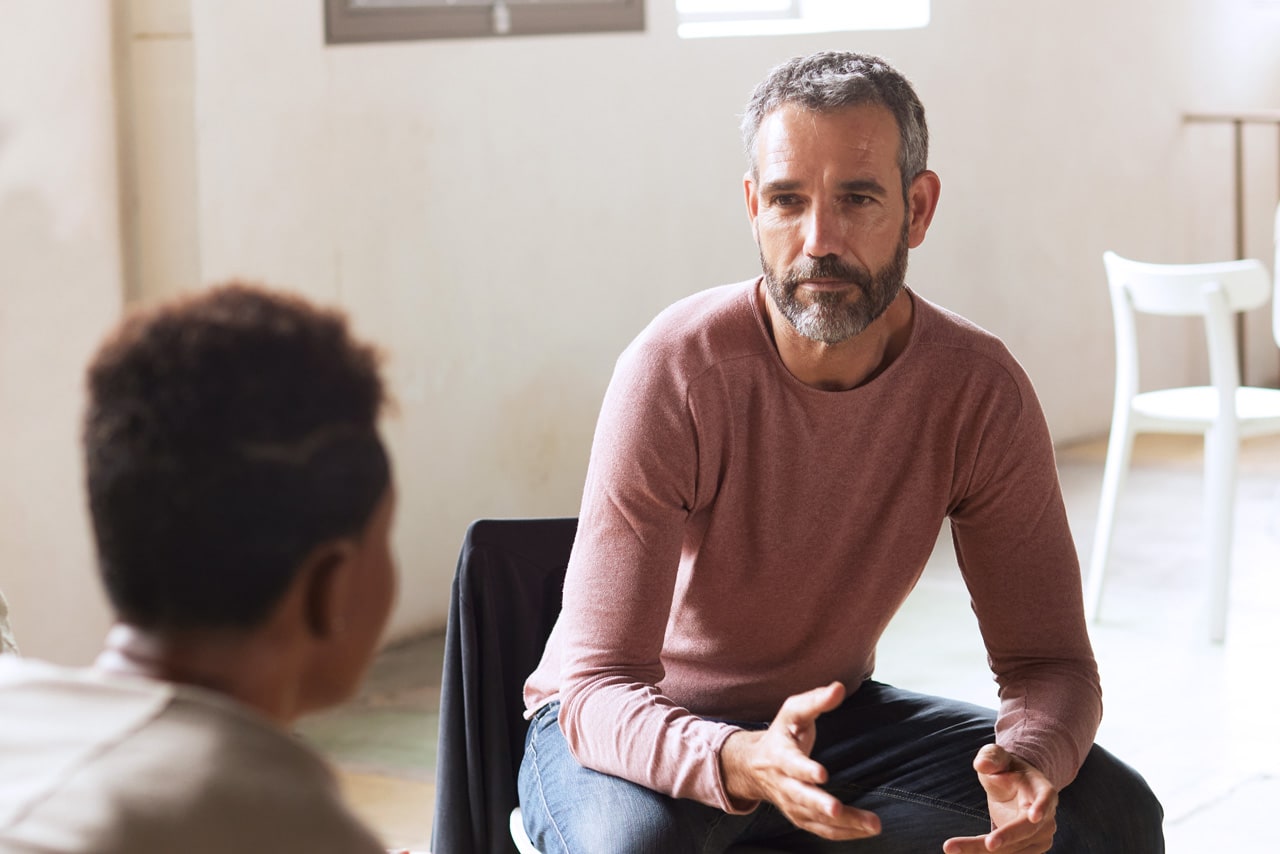 In einem hellen Raum hält ein sitzender mittelalter Mann erklärend die Hände offen.
