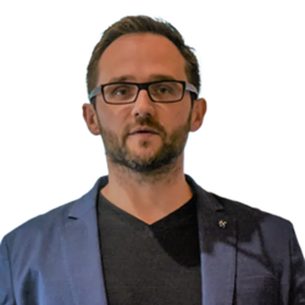 A man with glasses and a beard wearing a blue blazer over a black shirt, looking forward.