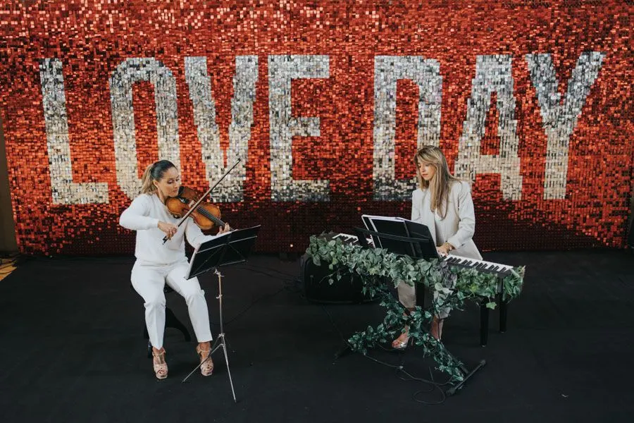 Dos mujeres músicas tocando el violín y el teclado en el escenario de la feria de bodas LOVE DAY Santander con un fondo brillante que forma la palabra 
