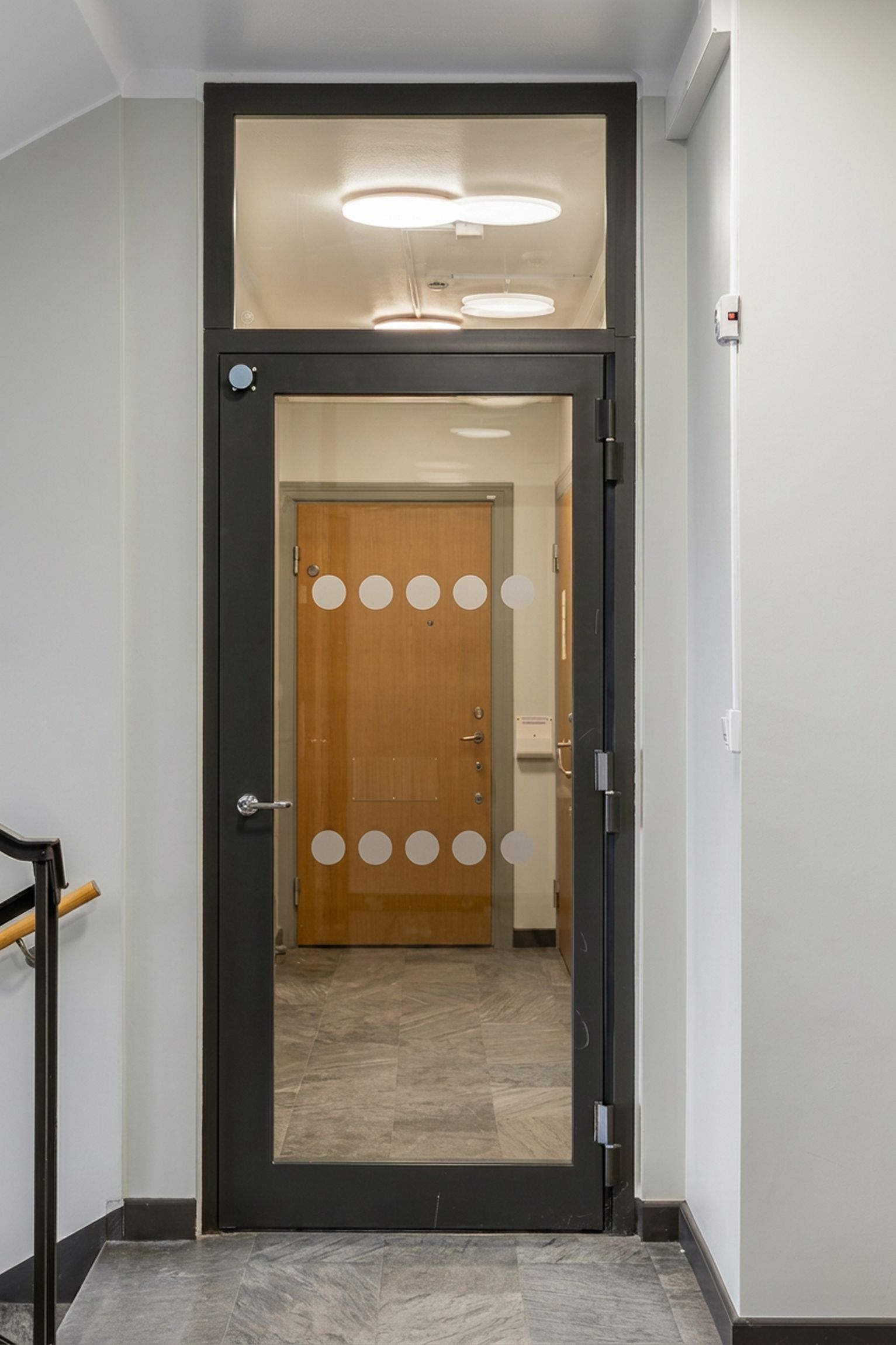 Glassdörr med vertikala rader av vita prickar leder in i en hall med träfärgad dörr i bakgrunden. Golvet är av grå marmor.