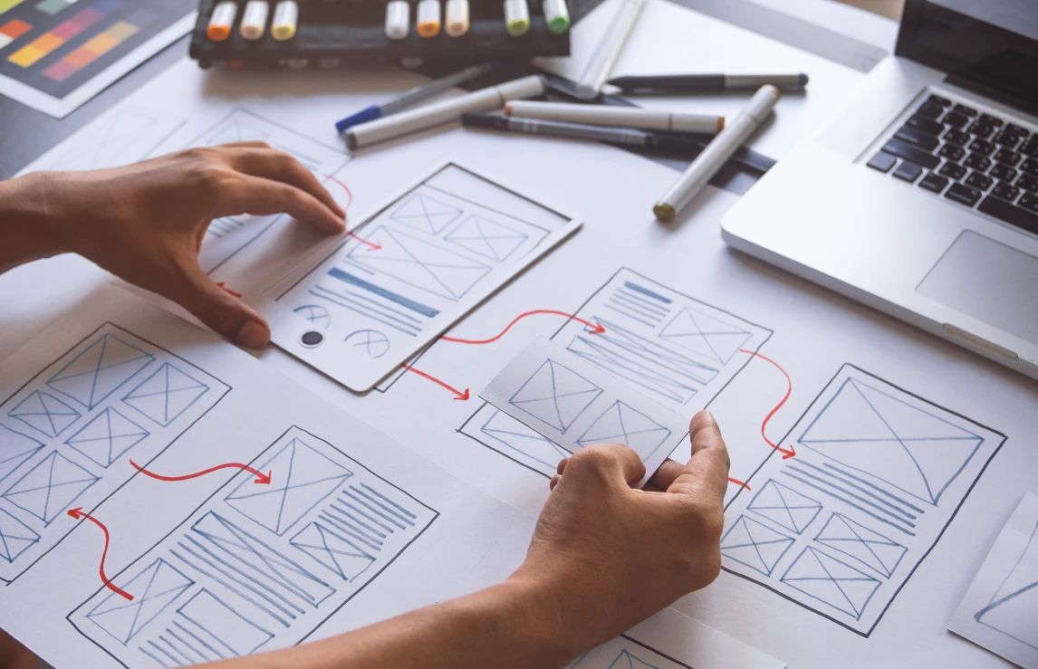 Hands arranging paper UX wireframe sketches connected by directional arrows, illustrating a website design process