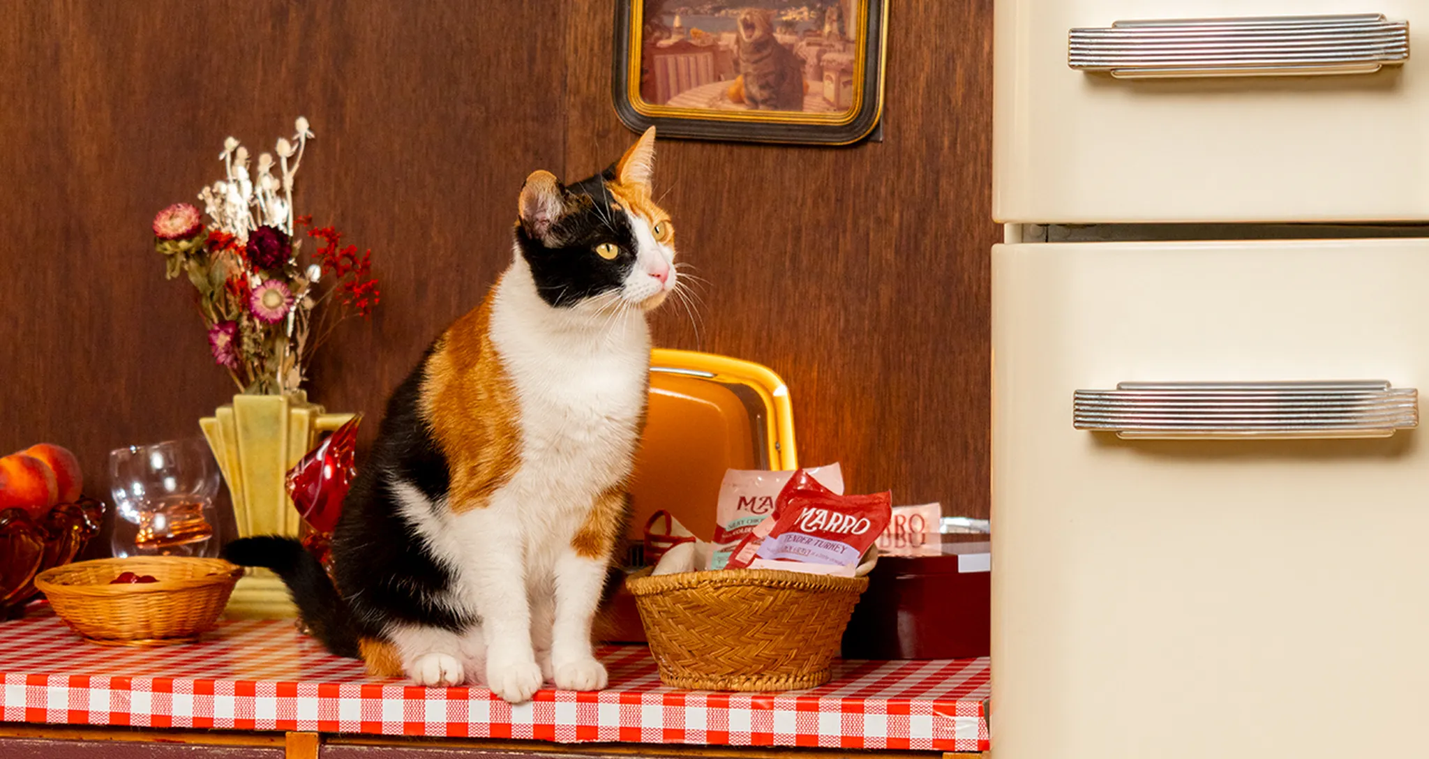 Calico cat sitting on a red-checkered tablecloth near a white fridge, surrounded by flowers and kitchen items.