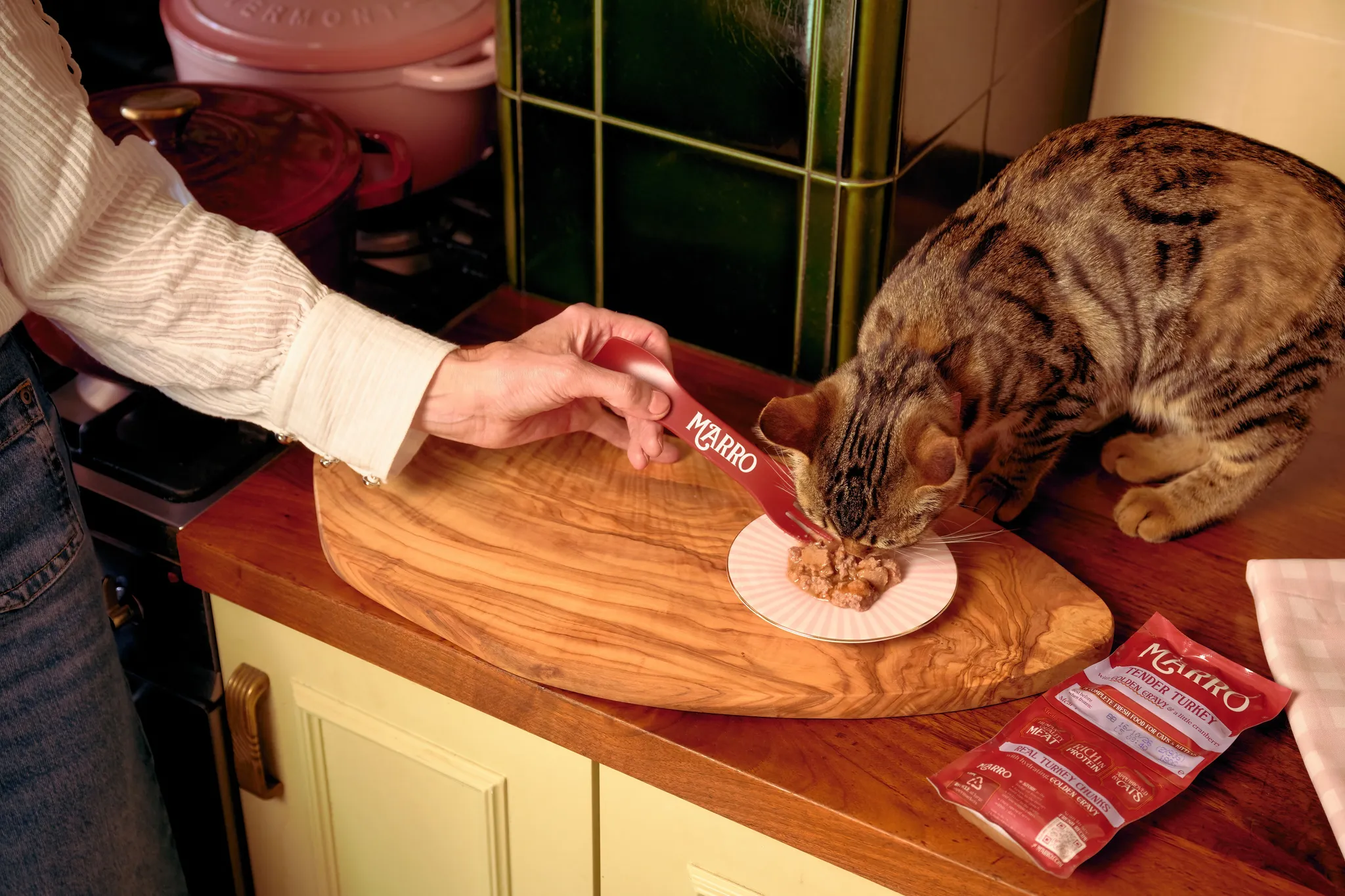 Cat eating from a plate on a wooden countertop, with a person using a utensil. Cat food packet labeled "Marro" beside them.