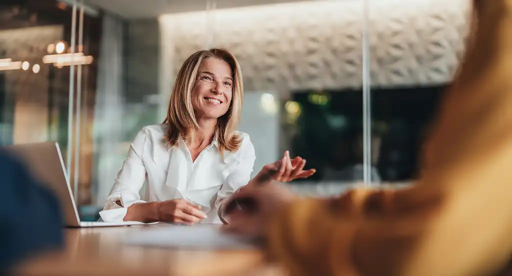 Smiling woman in a white blouse sits at a table during a meeting, gesturing, with a laptop open. Modern office setting in the background.