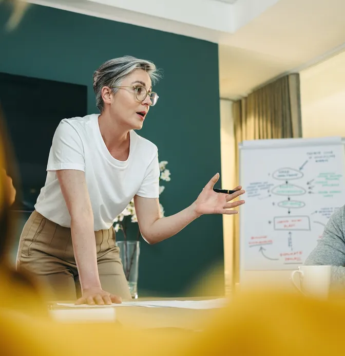Person with short hair and glasses leading a meeting, gesturing animatedly by a conference table and a whiteboard with diagrams in the background.