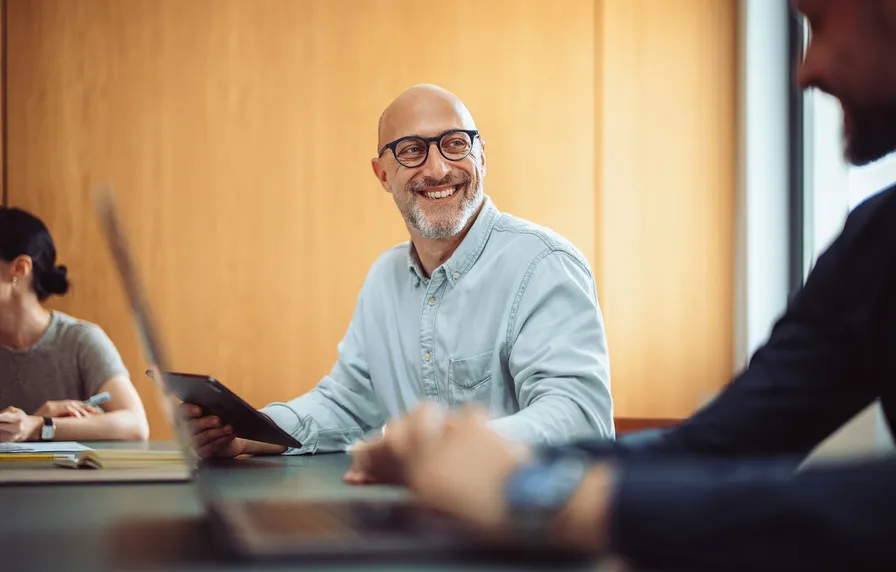 Smiling man with glasses holds a tablet in a meeting room, interacting with colleagues around a table with laptops.