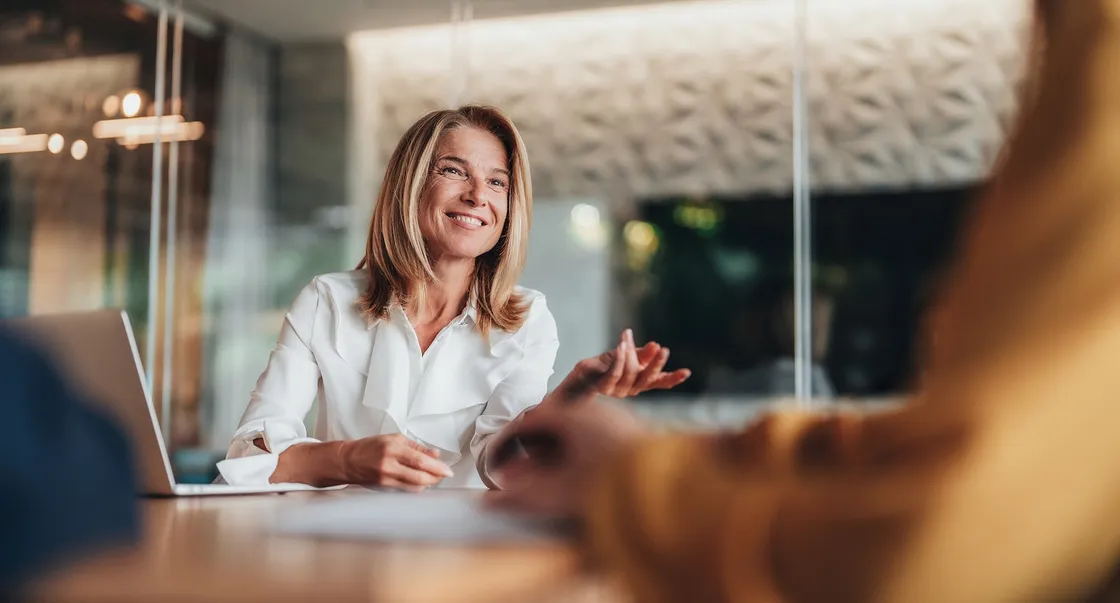 Smiling woman in a white blouse, engaged in conversation at a meeting table, with a laptop nearby and blurred figures in the foreground.