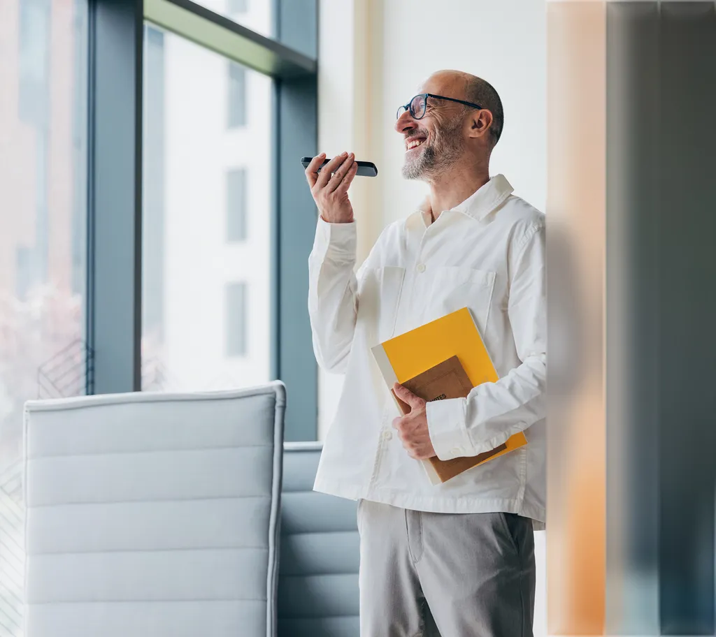 A man in a white shirt, holding a yellow folder, smiles while speaking into a smartphone, standing beside a window with soft light.