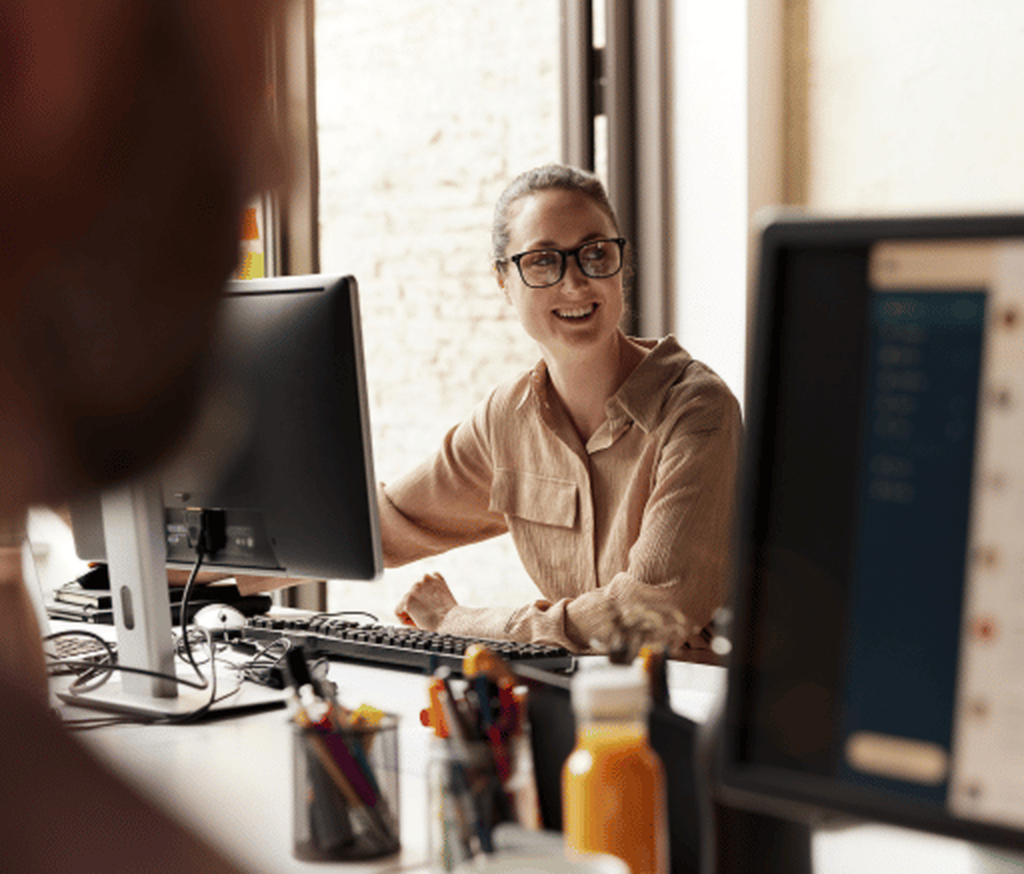 Lächelnde Frau am Arbeitsplatz sitzt am Schreibtisch vor PC-Monitor