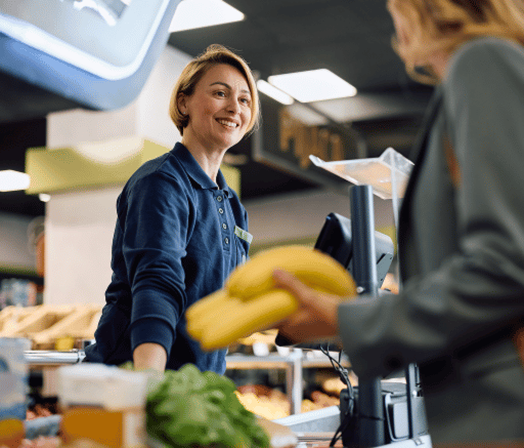 Frau am Kassierband lächelt Frau mit Bananen in der Hand an
