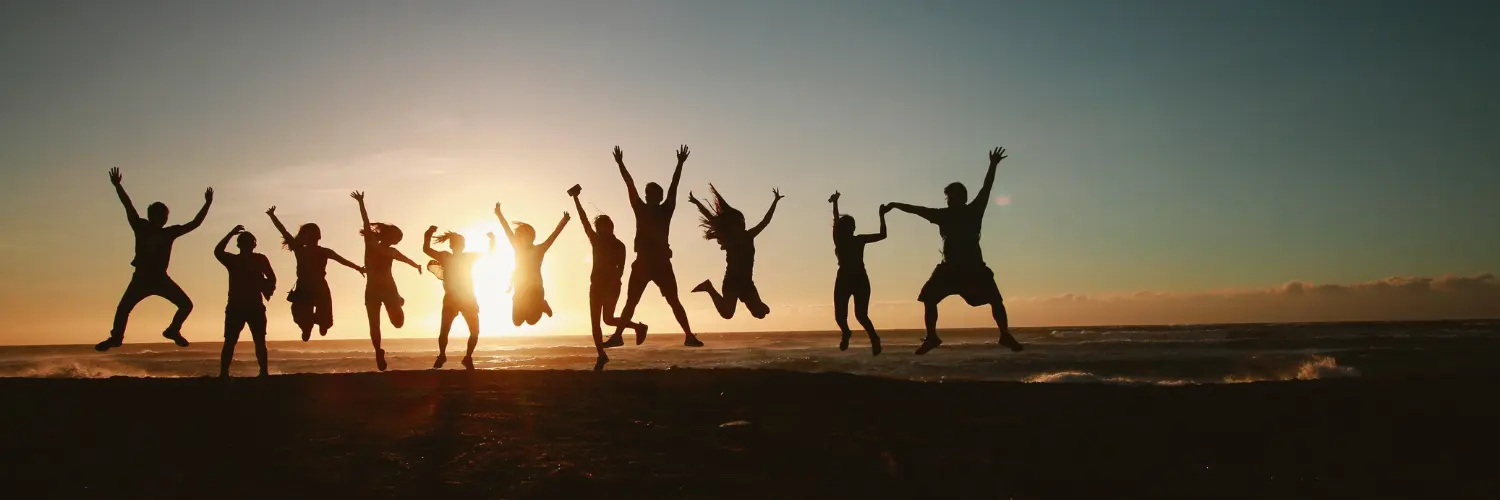 Silhouetten von Menschen, die bei Sonnenuntergang voller Freude am Strand springen, während die Sonne tief am Horizont steht und der Himmel darüber klar ist.