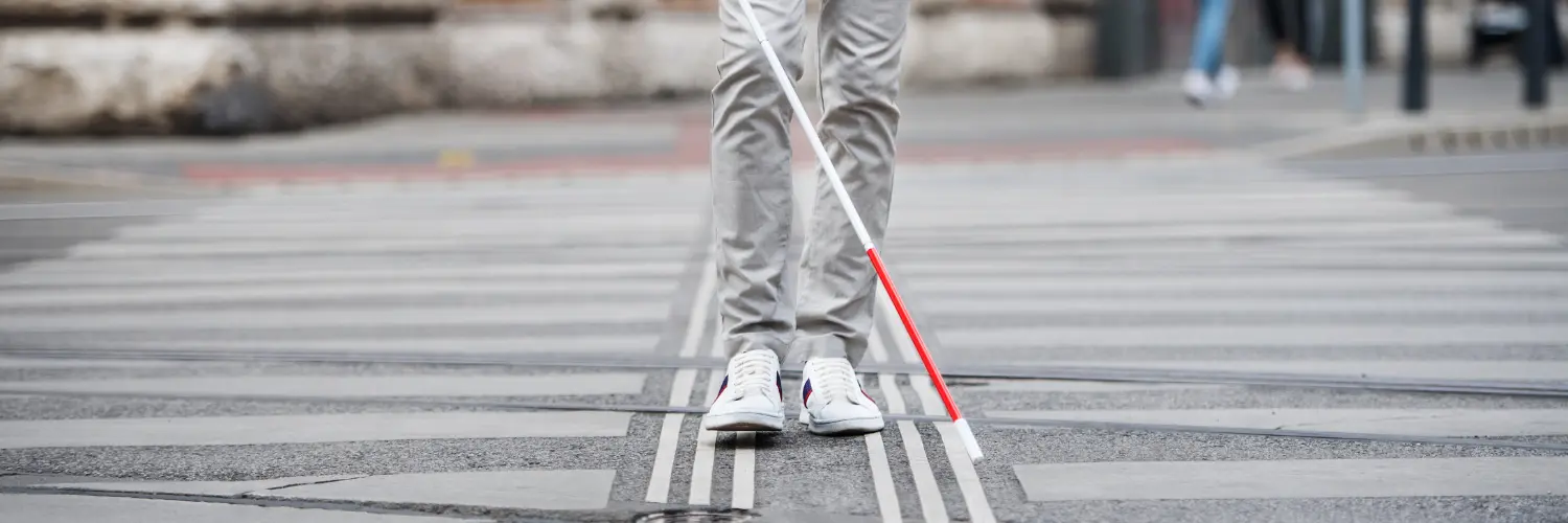 Eine Person mit einem Blindenstock geht über einen gestreiften Fußgängerüberweg, gekleidet in helle Kleidung und Sneaker.