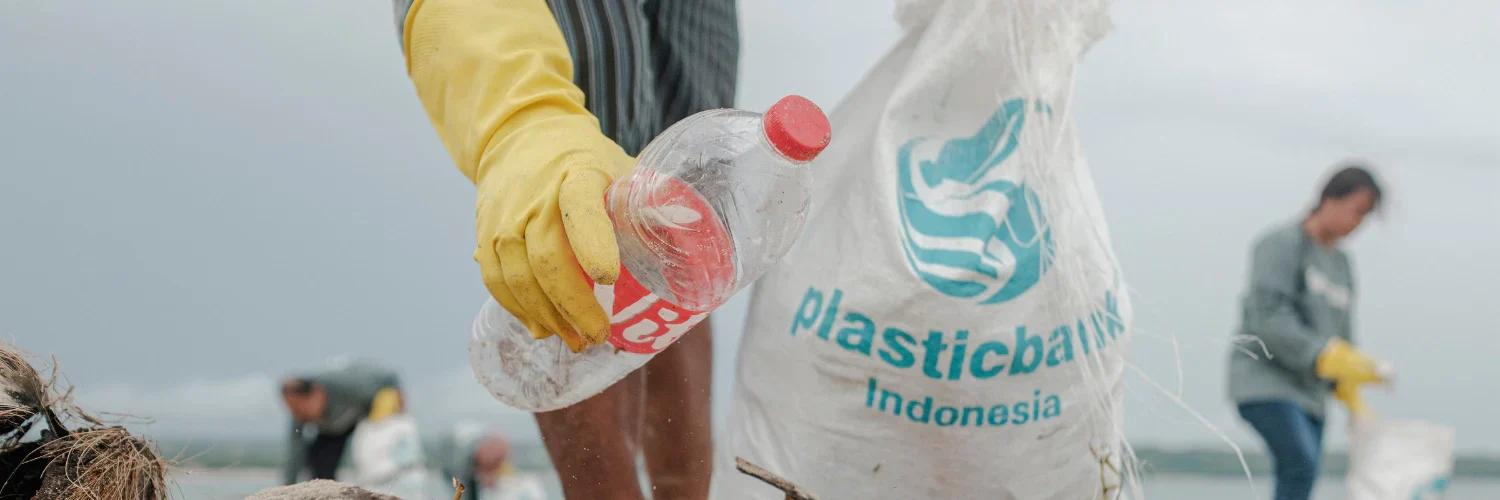 Eine Person mit gelben Handschuhen hebt eine Plastikflasche auf und legt sie während einer Aufräumaktion in eine Tasche mit der Aufschrift „plasticbank Indonesia“.