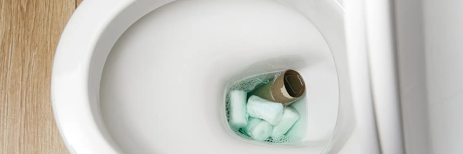 Toilettenbecken mit einem Kunststoffbehälter, der eine Papphülse und grüne Reinigungstabs im Inneren enthält.