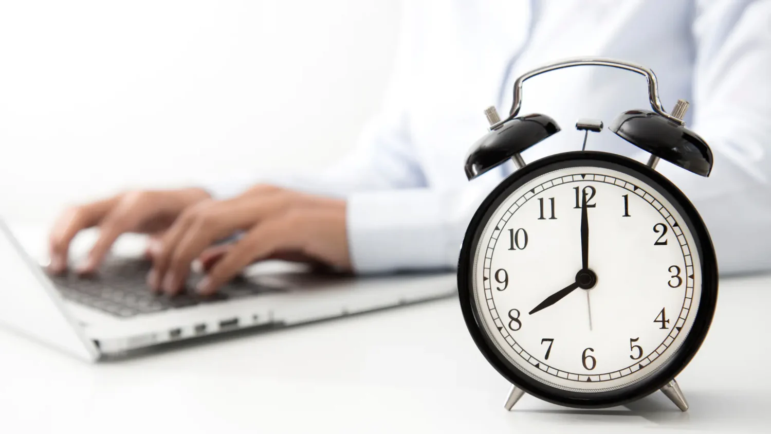 Ein klassischer Wecker im Vordergrund, der 7:00 Uhr anzeigt, während im Hintergrund Hände auf einem Laptop tippen.