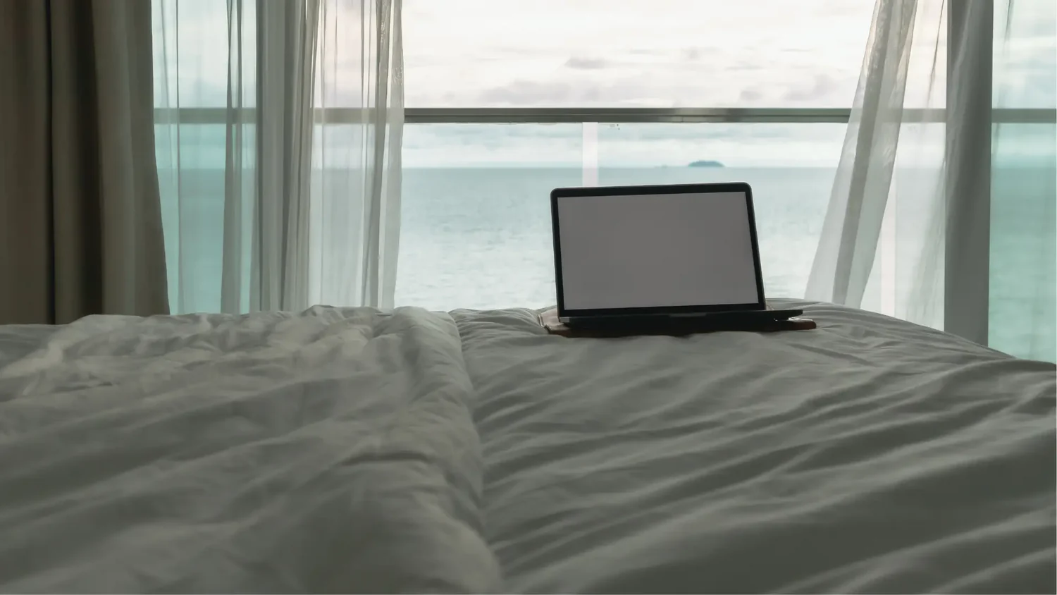 Ein geöffneter Laptop auf einem ordentlich gemachten Bett mit weißen Laken, mit Blick auf ein großes Fenster, das eine Aussicht auf das Meer unter einem bewölkten Himmel zeigt.
