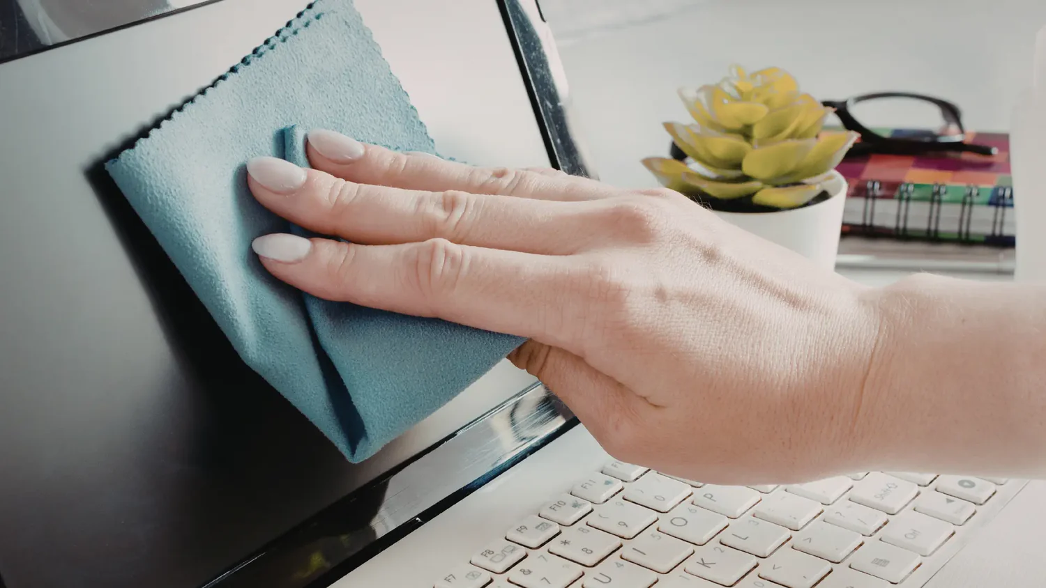 Eine Hand reinigt einen Computerbildschirm mit einem Mikrofasertuch; im Hintergrund sind eine kleine Pflanze und Notizbücher zu sehen.