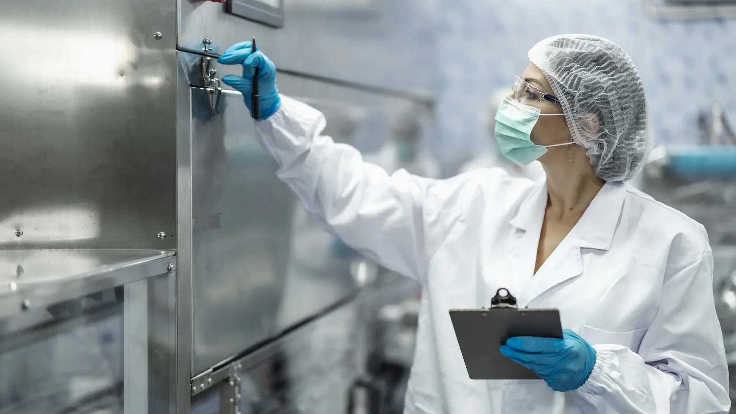 Ein Wissenschaftler im Laborkittel, mit Haarnetz und Maske, inspiziert die Ausrüstung, während er ein Klemmbrett hält und mit behandschuhten Händen einen Regler einstellt.