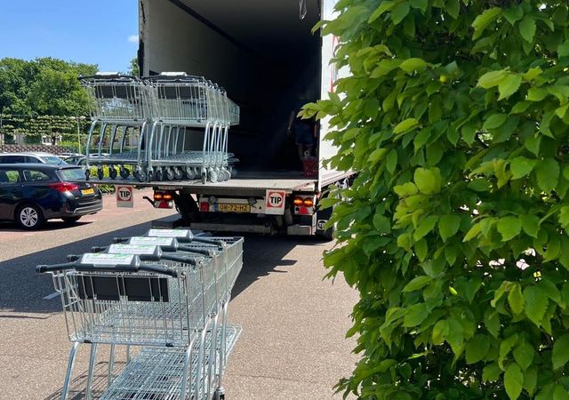 Winkelwagentjes worden op een zonnige dag uit een vrachtwagen gelost op een parkeerplaats, met groen op de voorgrond.
