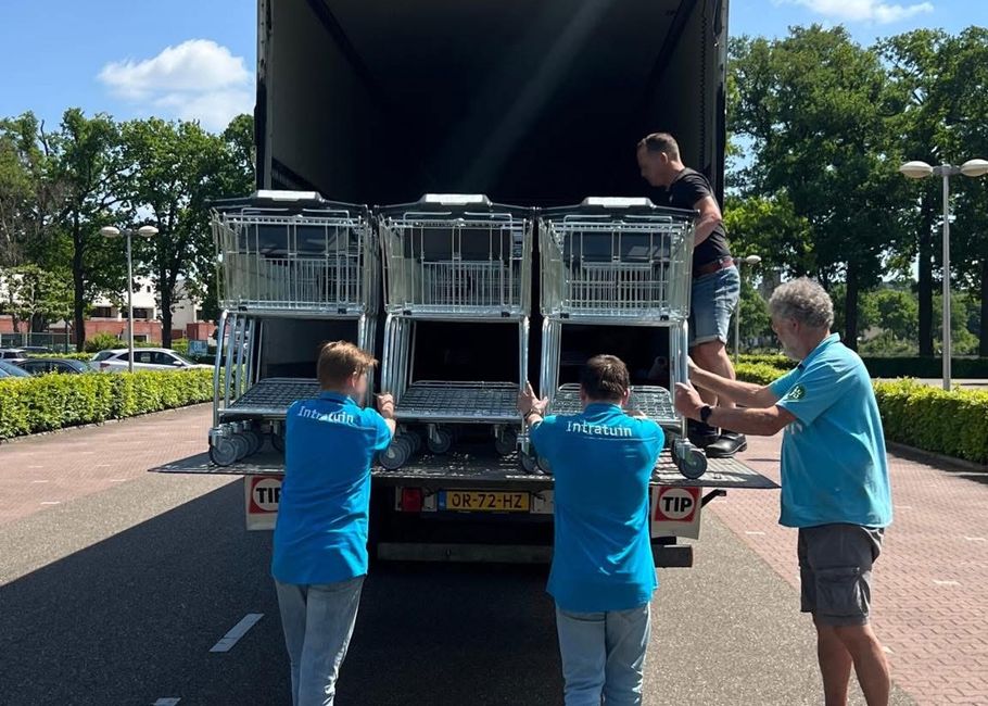 Twee mannen in blauwe shirts en twee anderen laden op een zonnige dag winkelwagentjes uit een vrachtwagen, omringd door bomen en geparkeerde auto's.