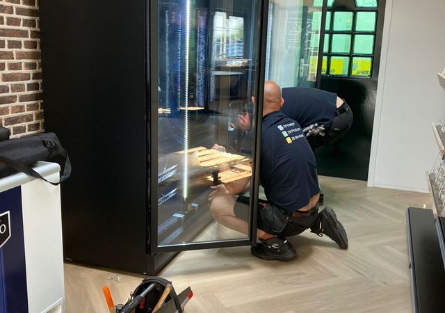 Twee arbeiders installeren een grote zwarte vitrinekoelkast in een kamer met bakstenen muren en houten vloeren. Er ligt een gereedschapstas vlakbij op de grond.
