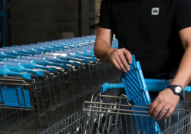 Een persoon in een zwart shirt hanteert een rij winkelwagentjes met blauwe handvatten in een magazijnomgeving.