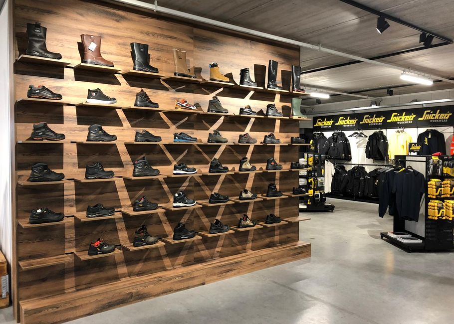Winkelinterieur met een houten wand vol planken waarop laarzen en schoenen staan uitgestald; daarnaast rekken met zwart-gele werkkleding.