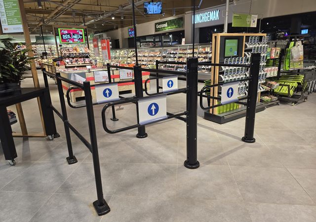 Ingang van een supermarkt met een zwarte tourniquetpoort, borden met blauwe pijlen en schappen gevuld met verschillende producten op de achtergrond.