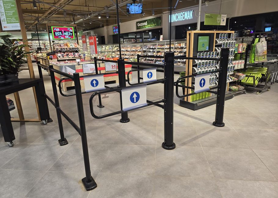 Ingang van een supermarkt met een zwarte tourniquetpoort, borden met blauwe pijlen en schappen gevuld met verschillende producten op de achtergrond.