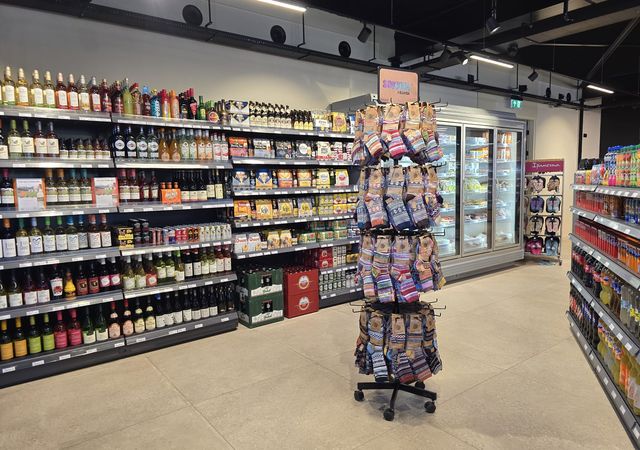 Een gangpad in de supermarkt met schappen met wijn, drankjes, snacks en een wisselende uitstalling van chips. Op de achtergrond staat een koelkast met drankjes.