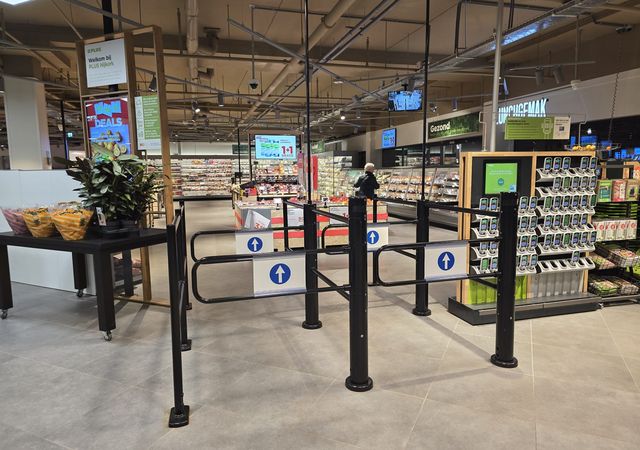 Ingang van de supermarkt met zwarte eenrichtingsdraaipoorten gemarkeerd met blauwe pijlen, gangpaden, displays en een shopper op de achtergrond.