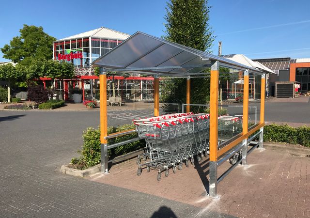 Winkelwagenopvang met verschillende karren erin, gelegen op een parkeerplaats bij een winkel met een rood bord op de achtergrond.