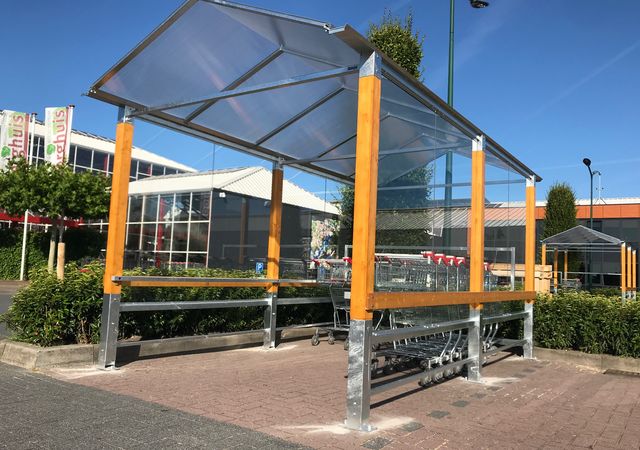 Winkelwagenafdak met transparant dak en houten steunen, gelegen op een parkeerplaats onder een helderblauwe hemel.