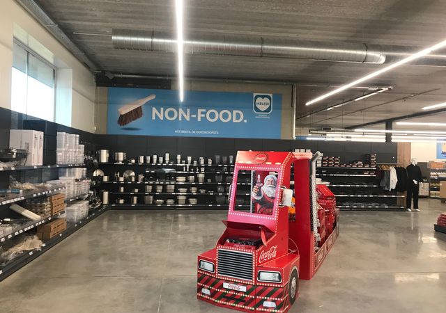 Een winkelpad met het opschrift 'Non-Food' met planken met keukengerei en een display met rood Coca-Cola-thema dat lijkt op een vrachtwagen.