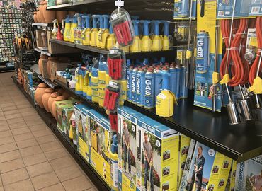 Winkelgang met tuingereedschap, gasflessen en besproeiingsaccessoires op de planken. Op de achtergrond zijn kleipotten zichtbaar.