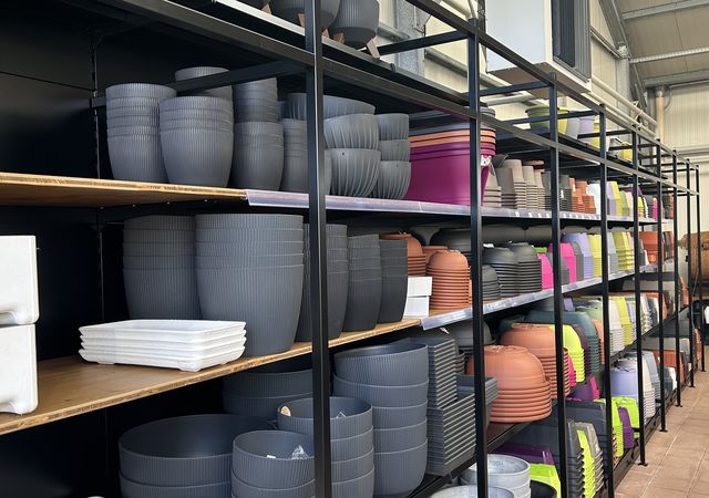 Planken gevuld met verschillende maten en kleuren plantenpotten, waaronder grijs, wit, oranje en veelkleurig, in een winkelomgeving.