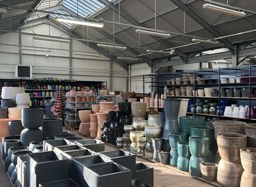 Een ruim magazijn met rijen gestapelde keramische potten in verschillende vormen en kleuren, onder een hoog plafond met dakraam.
