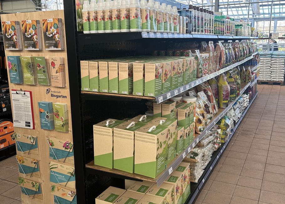 Een winkelgang met planken vol tuinbenodigdheden, waaronder grondzakken, zaadpakketten en plantenverzorgingsproducten, onder fel licht.