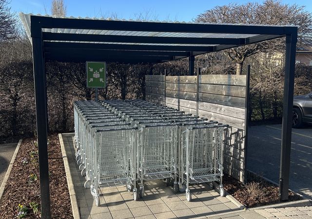 Een overdekte buitenruimte met netjes gestapelde winkelwagens op een verharde ondergrond, omgeven door een laag houten hekwerk en mulch.