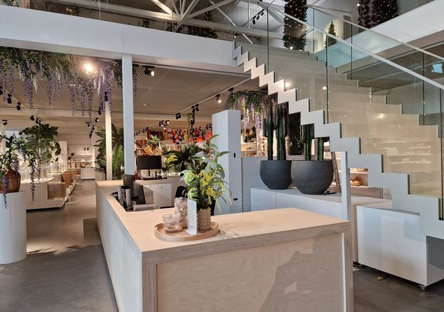Modern interieur van een winkel met een houten toonbank, glazen trap en diverse planten en bloemen tentoongesteld.