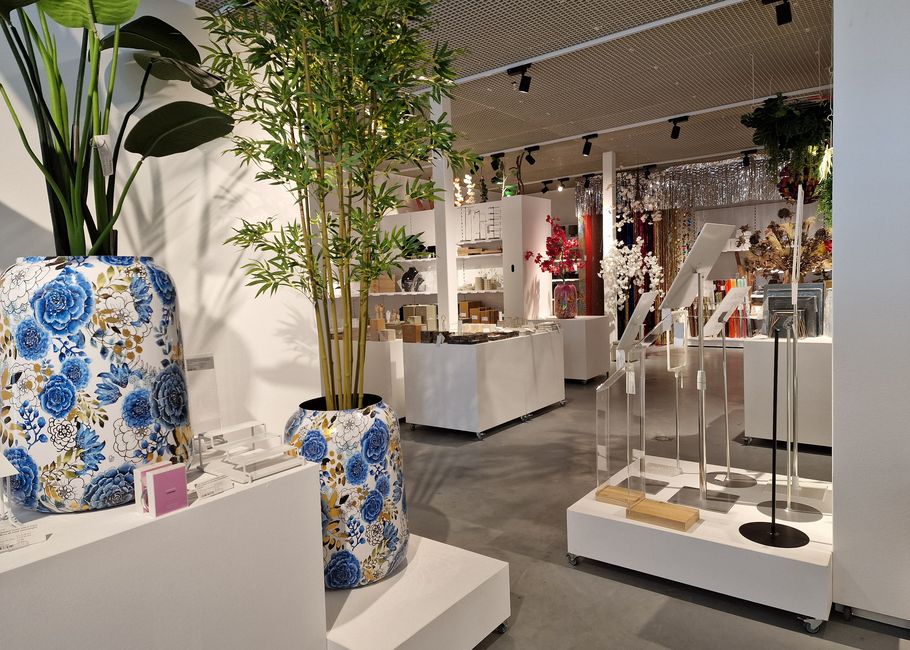 Een modern winkelinterieur met grote bloemenvazen, planten en displays van diverse decoratieve artikelen op witte planken en stands.