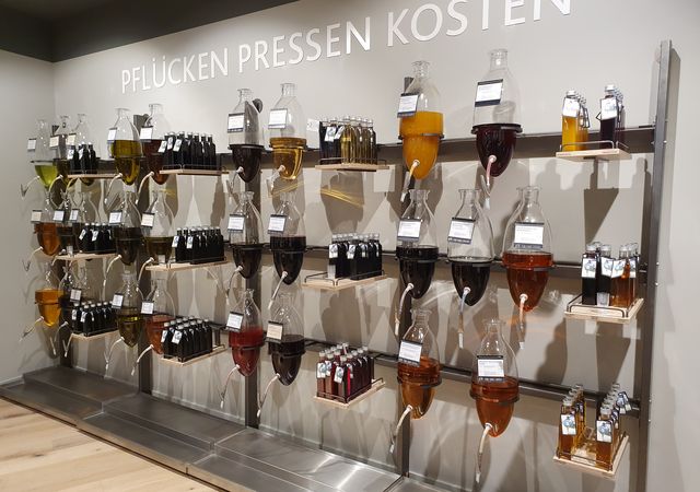 Wanddisplay met glazen dispensers en flessen met diverse amberkleurige en gele vloeistoffen op planken onder het bord "PFLÜCKEN PRESSEN KOSTEN".